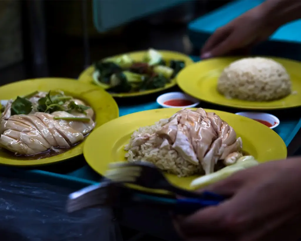A close-up of Hainanese chicken rice on yellow plates; includes sliced chicken, rice, cucumbers, and greens. A hand serves the meal with dipping sauces.
