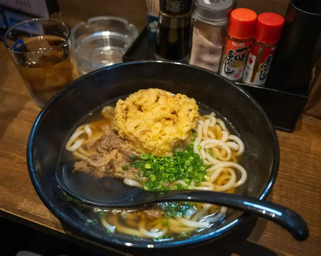 A bowl of udon noodles in broth is topped with golden tempura and chopped green onions, alongside beef. Nearby are seasonings and a glass of iced tea.