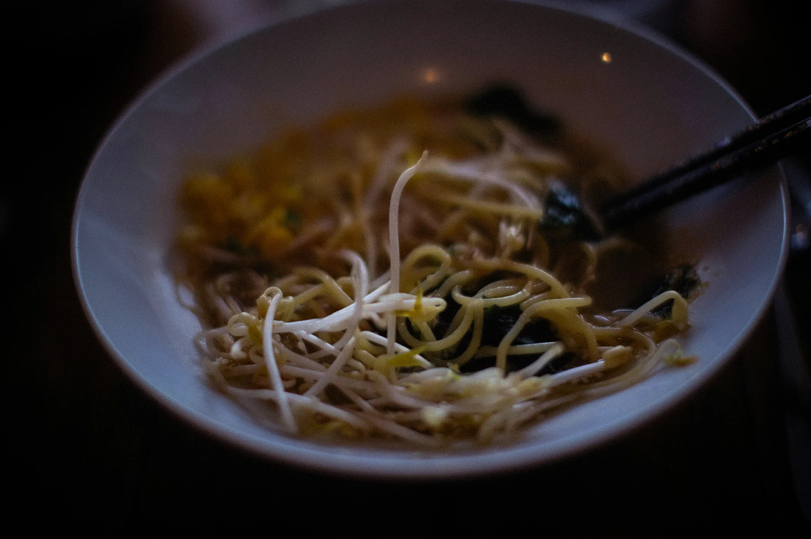 Close-up of a bowl of Asian noodle soup with bean sprouts, chopsticks, and dark moody lighting in a cozy restaurant setting