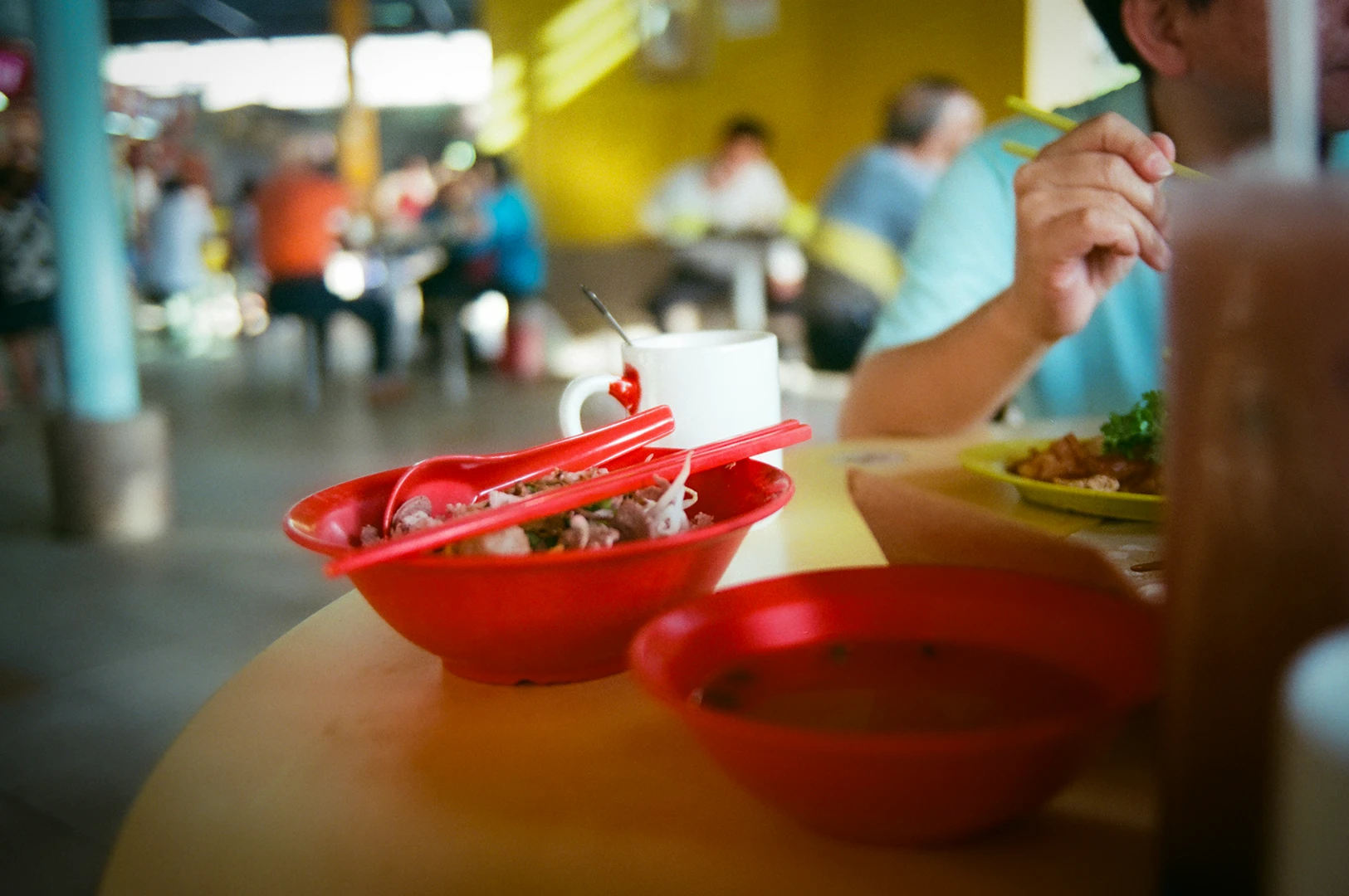 Close-up of a hawker centre table in Singapore with red bowls of food, chopsticks, and a person eating in a casual dining setting.