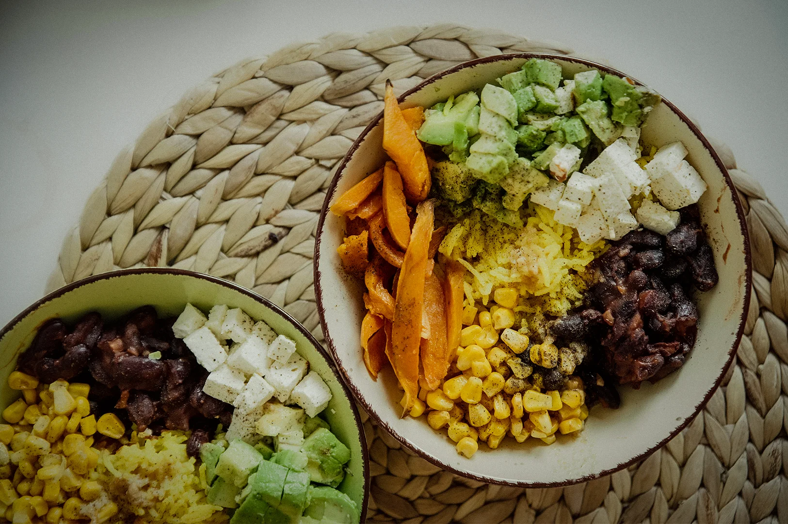 Healthy vegan buddha bowls with avocado, black beans, sweet potatoes, corn, rice, and tofu served in ceramic bowls on a woven placemat table setting