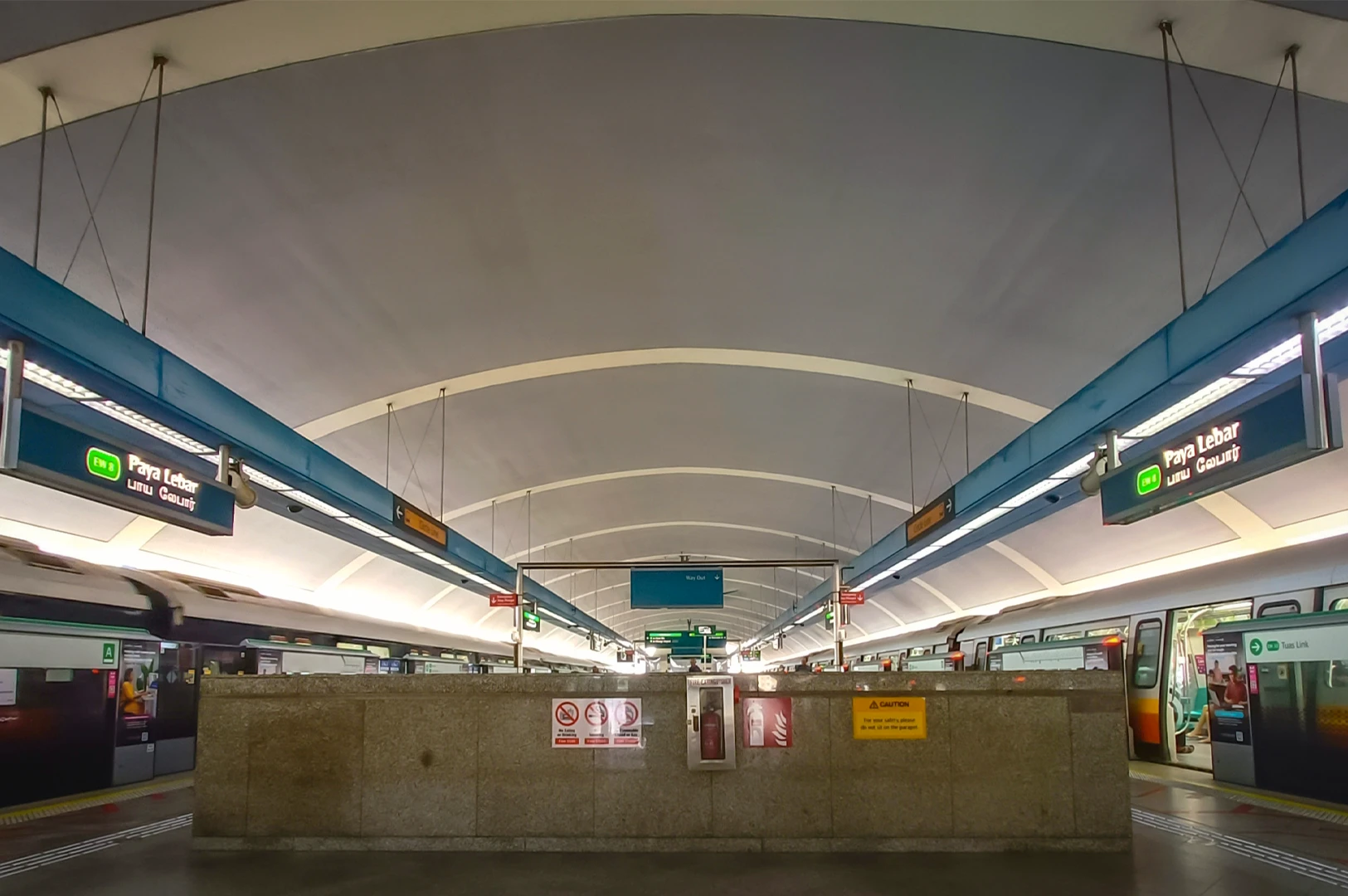 Wide angle view of Paya Lebar MRT station platform in Singapore with trains on both sides, overhead route signs and curved modern station architecture
