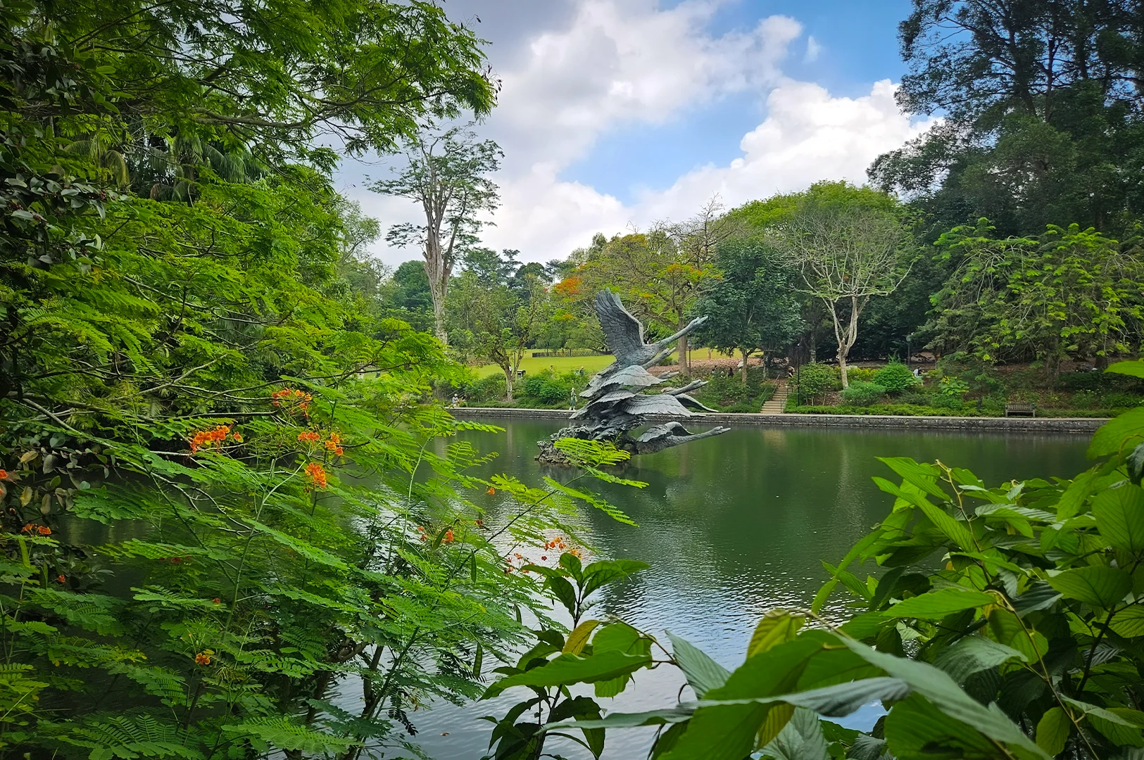Lush greenery surrounding a calm lake at Singapore Botanic Gardens, featuring a dynamic bird sculpture rising from the water with trees and flowering plants in the foreground.