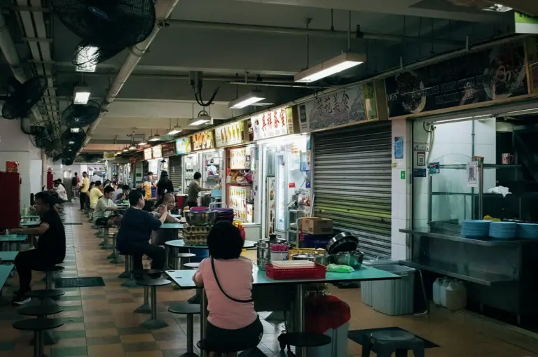 Interior of a Singapore hawker centre with diners seated at communal tables and local food stalls lining the walkway under fluorescent lighting