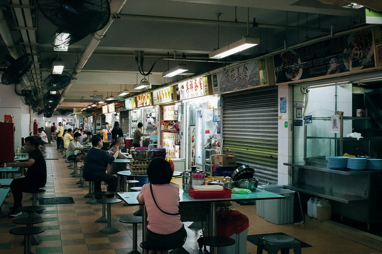 Interior of a Singapore hawker centre with diners seated at communal tables and local food stalls lining the walkway under fluorescent lighting