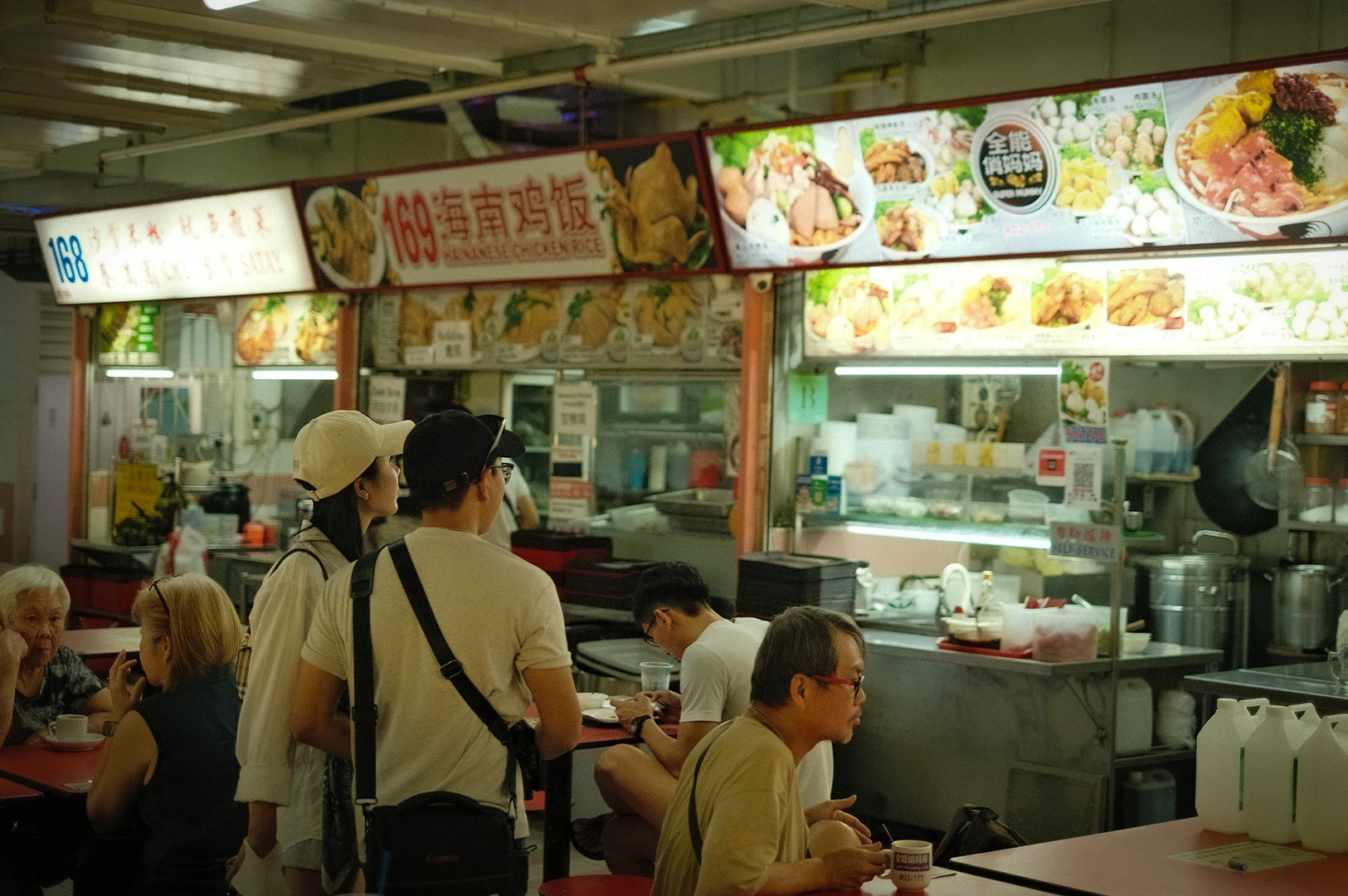 Customers ordering at a busy hawker stall serving Hainanese chicken rice, with bright menu boards and diners seated at communal tables.