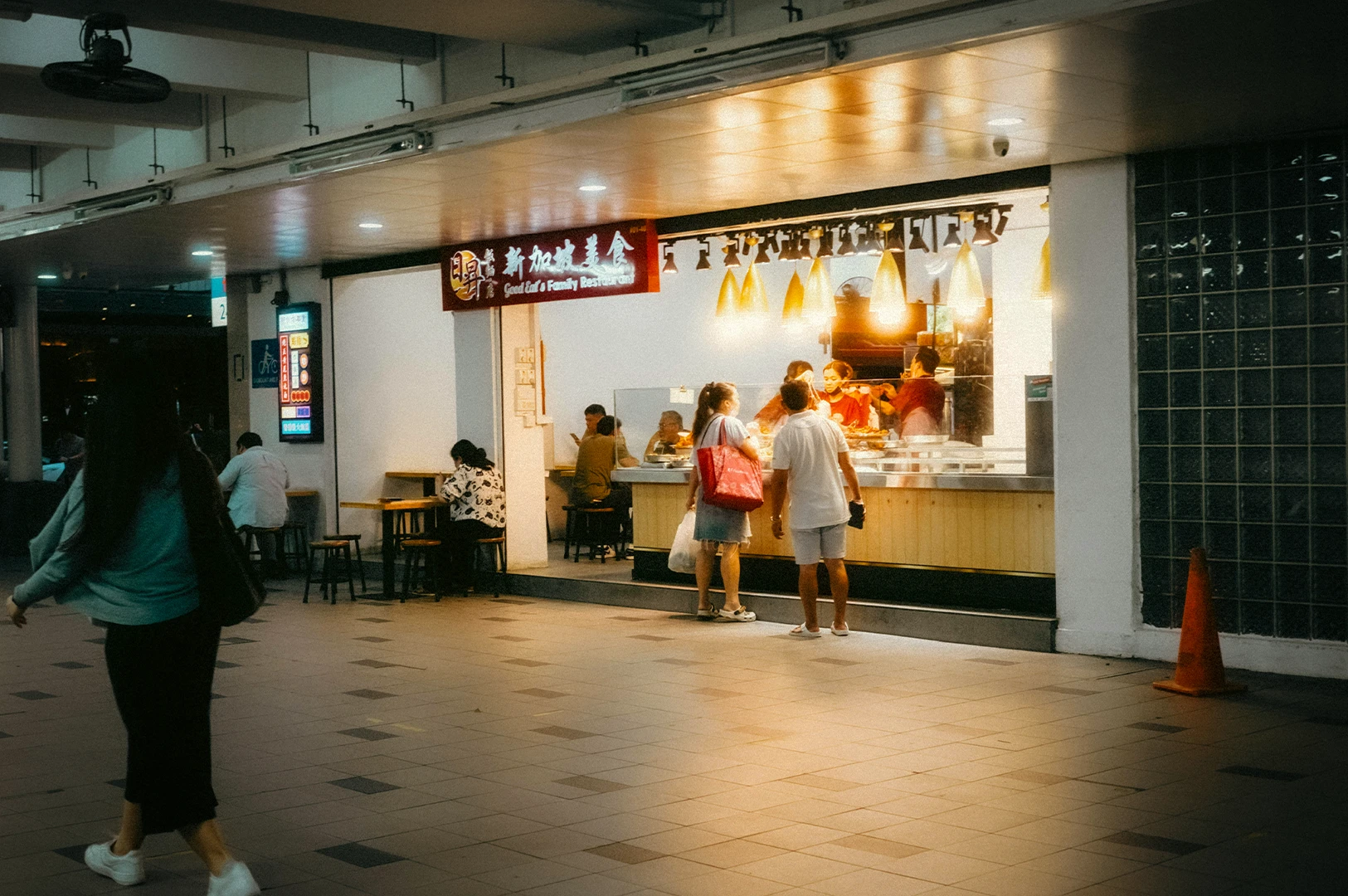 People ordering food at a brightly lit hawker stall in Singapore at night, with diners seated and walking through a covered food court.