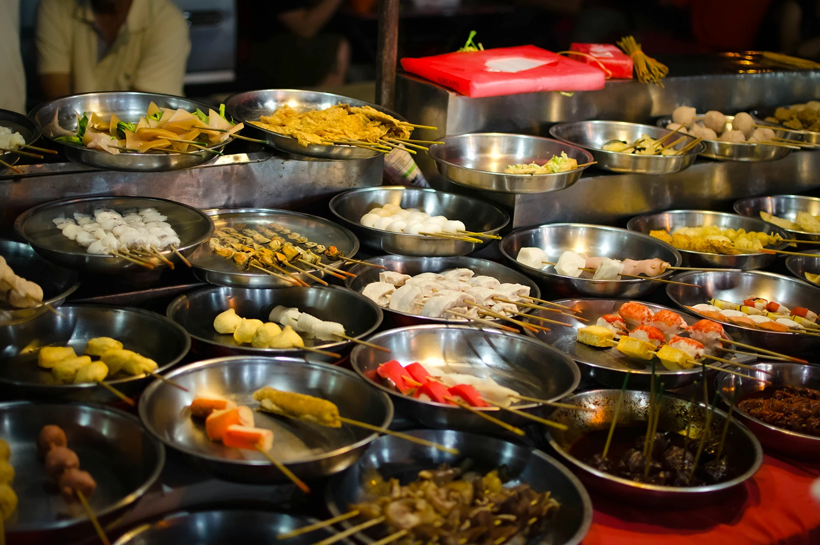 Variety of street food skewers including seafood, meat, and vegetables displayed in metal trays at a night market stall.
