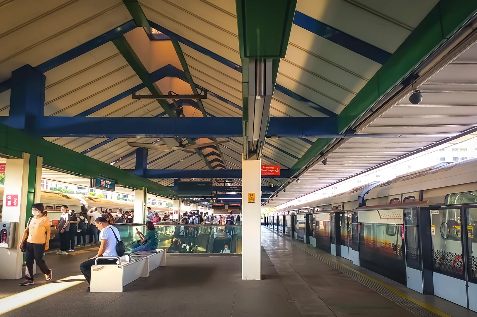Commuters waiting on the elevated outdoor platform of Yishun MRT station in Singapore, featuring the iconic green and blue pitched roof architecture and a parked North-South Line train.