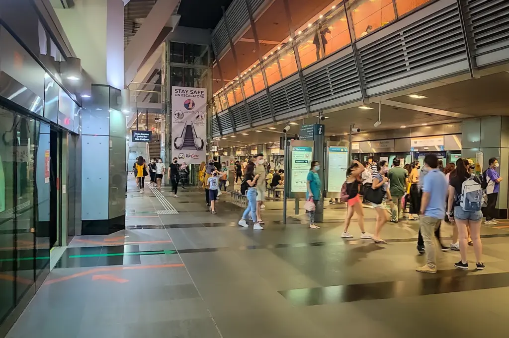 Busy concourse area inside Paya Lebar MRT station in Singapore with commuters walking near train platforms, escalators and illuminated upper level walkway at night