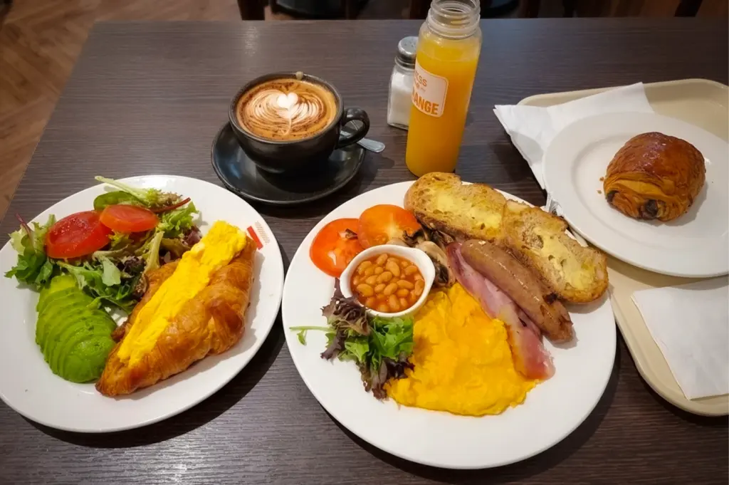 Brunch spread at The Sandwich Shop Cluny Court featuring scrambled eggs with sausage and bacon, croissant with eggs and avocado salad, latte with heart latte art, orange juice, and a chocolate pastry.
