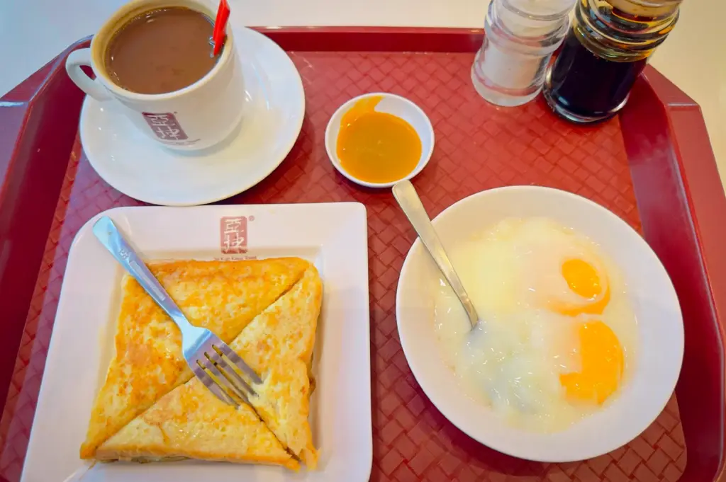 A traditional Ya Kun Kaya Toast set on a red tray, featuring thin-sliced toasted bread, two soft-boiled eggs in a bowl, and a cup of hot Nanyang-style kopi.