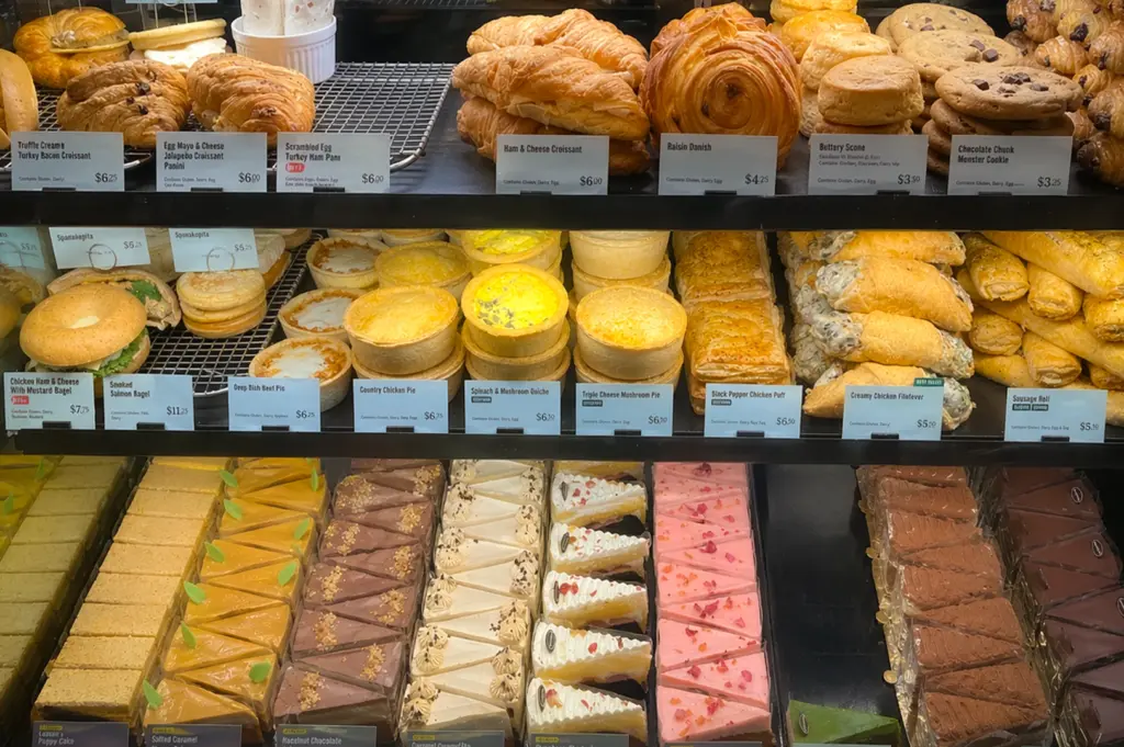 A well-lit multi-tier display case at Starbucks Northpoint City, Singapore, featuring an array of fresh pastries and cakes. The top shelf holds croissants, raisin danishes, scones, and chocolate cookies. The middle shelf displays savory items like quiches, meat pies, chicken puffs, and bagels. The bottom shelf showcases rows of colorful sliced cakes, including salted caramel, hazelnut chocolate, and strawberry flavors, all neatly arranged with visible price tags.