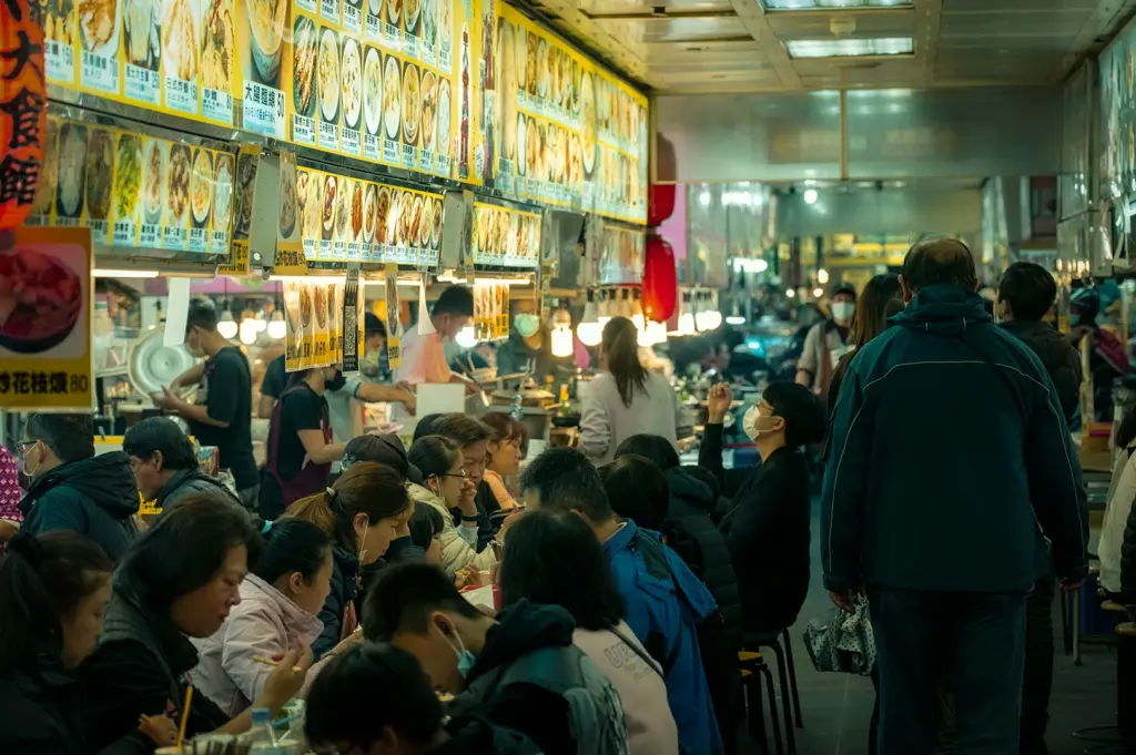 Crowded night market food stall with diners seated shoulder to shoulder, illuminated menu boards overhead, and cooks preparing dishes in a lively indoor alley.