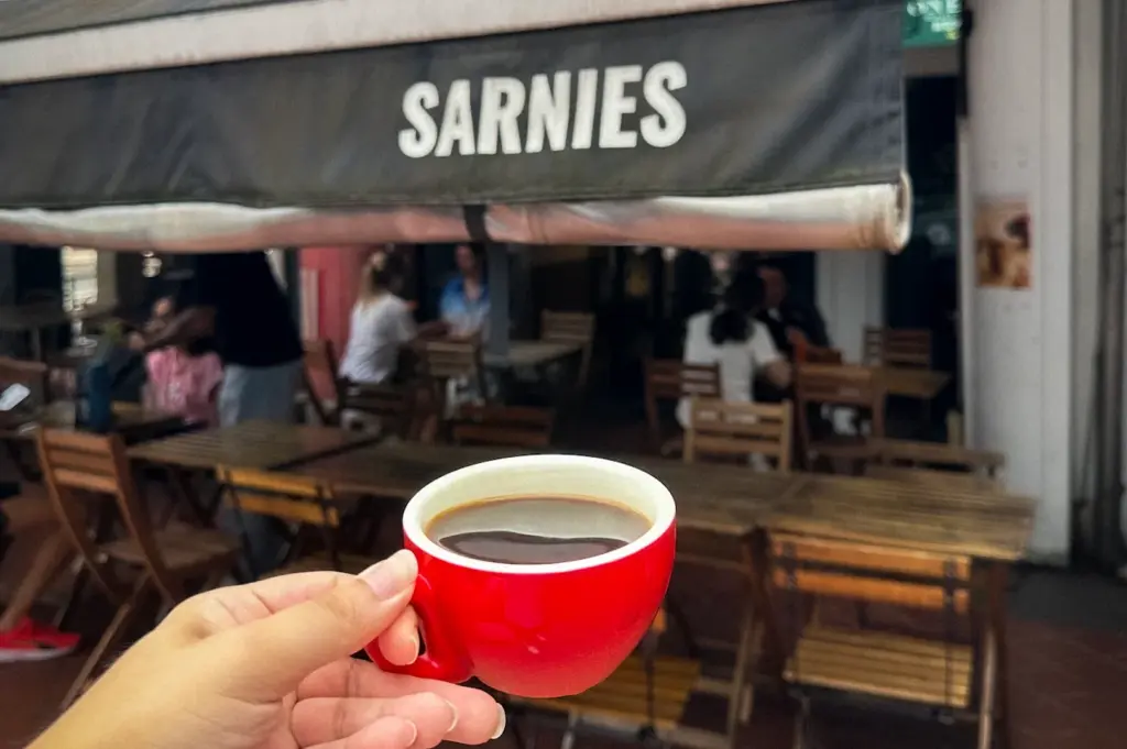 Hand holding a red cup of black coffee outside Sarnies café in Singapore with outdoor wooden seating and café awning in the background