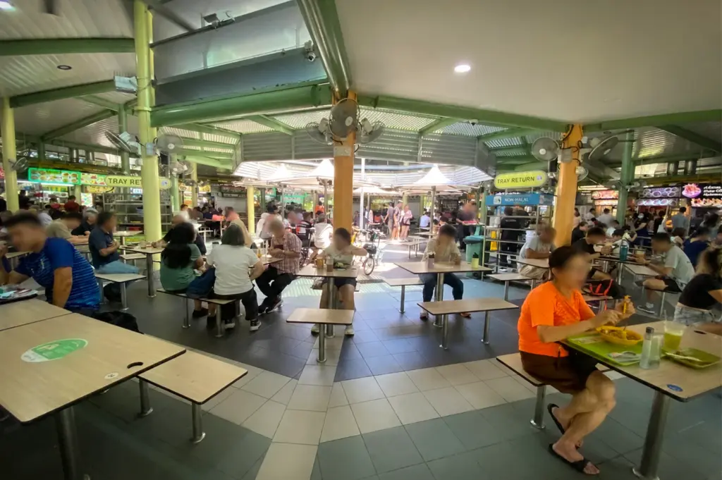 Busy Singapore hawker centre with diners seated at shared tables under a covered food court, surrounded by food stalls and tray return stations.