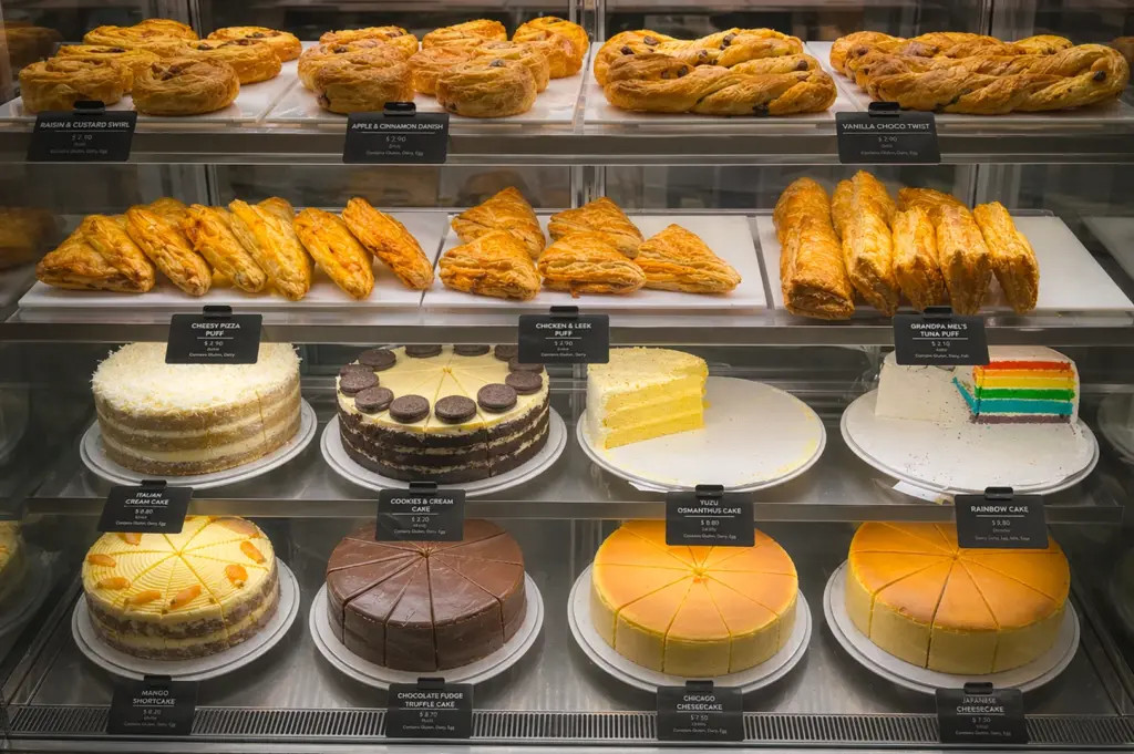 Pastry display at Coffee Bean & Tea Leaf Northpoint Singapore featuring danishes, puffs, and whole cakes like Chicago Cheesecake, Rainbow Cake, and Chocolate Fudge Truffle on tiered glass shelves.