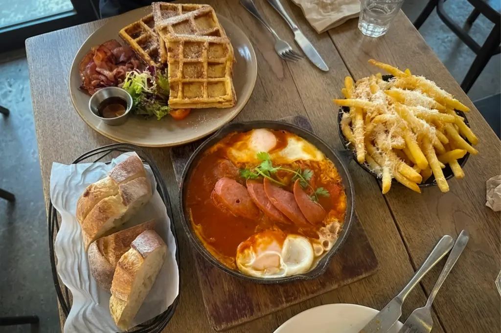 Hearty brunch at Nassim Hill Bakery Bistro featuring shakshuka with eggs and sausage, waffles with bacon, parmesan fries, and crusty bread on a wooden table.