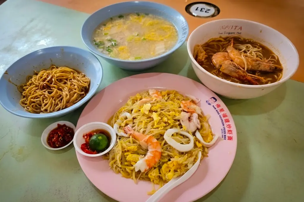 **File name:** singapore-hawker-hokkien-mee-prawn-noodles-meal **Alt text:** Singapore hawker meal with Hokkien mee on a pink plate, prawn noodle soup, dry noodles, egg drop soup, and chili lime dipping sauce on a table.