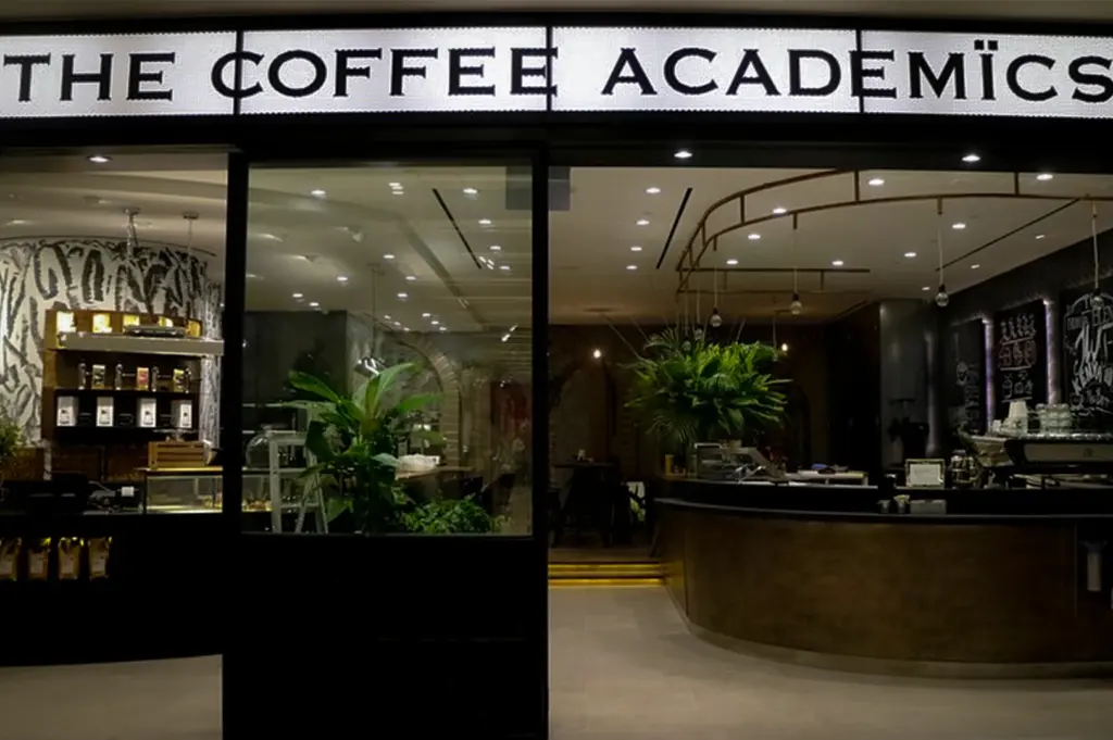 Front entrance of The Coffee Academics Singapore featuring a modern specialty coffee cafe interior with minimalist black decor, coffee bar counter, indoor plants and illuminated storefront signage