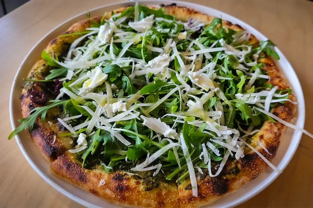 Vegetarian pizza at Real Food Grocer Singapore topped with fresh arugula, shaved cheese, olives, and pesto sauce on a crispy wood-fired crust served on a wooden table