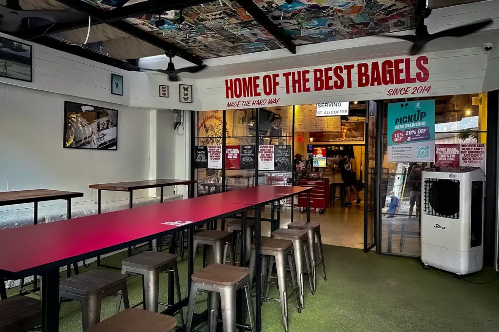 Outdoor seating area at Two Men Bagel House Singapore featuring communal tables, industrial-style stools, retro ceiling artwork and bold Home of the Best Bagels signage beside the cafe entrance