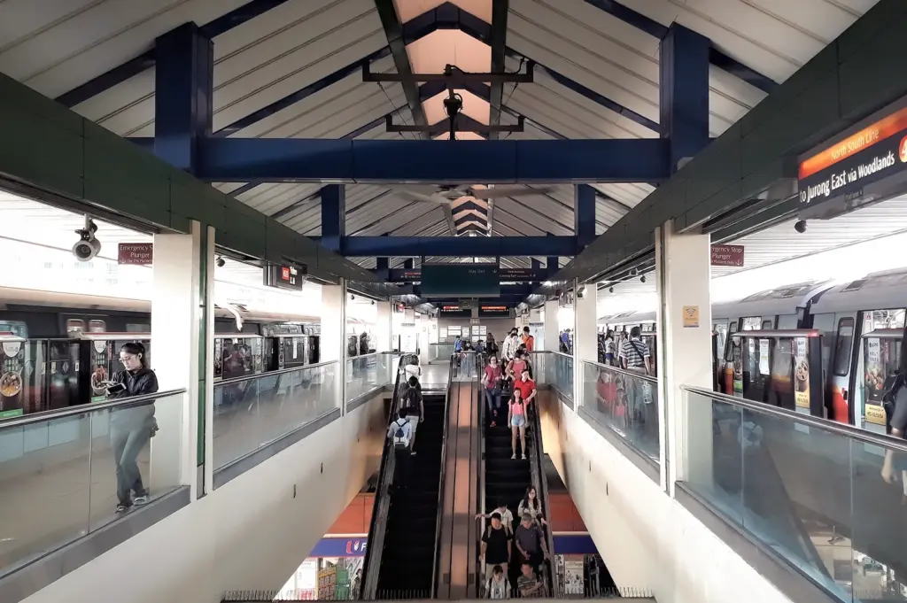 Bustling Yishun MRT Station platform in Singapore with commuters on escalators. North-South Line trains are visible at the platforms under a high vaulted ceiling with industrial blue beams.