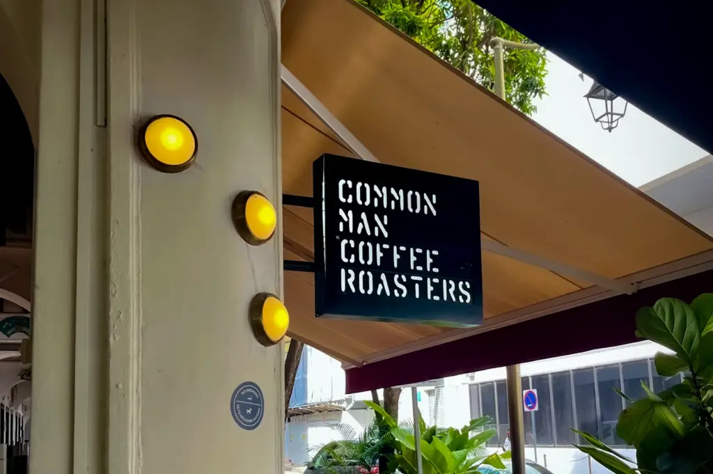 Outdoor seating area at Common Man Coffee Roasters Singapore with cafe tables, greenery, shaded awnings and illuminated hanging sign along a heritage shophouse walkway
