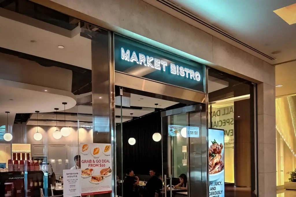 Modern entrance of Market Bistro restaurant in Singapore featuring illuminated signage, glass doors, and elegant indoor dining area inside a shopping mall corridor