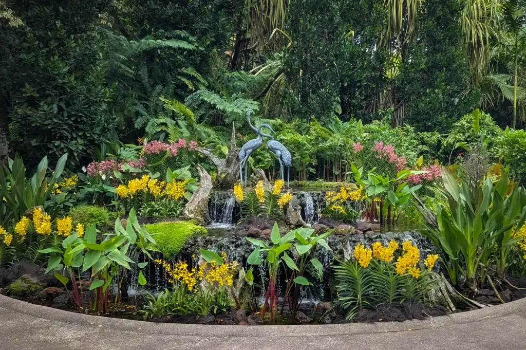 Lush tropical garden display at Singapore Botanic Gardens featuring cascading waterfalls, vibrant orchids, and a crane sculpture surrounded by dense greenery.