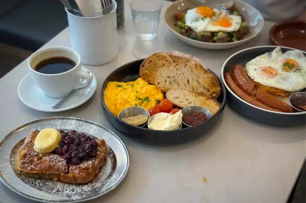 Atlas Coffeehouse Singapore brunch with scrambled eggs and sourdough, sausages with fried eggs, French toast with berries, and black coffee on a table.