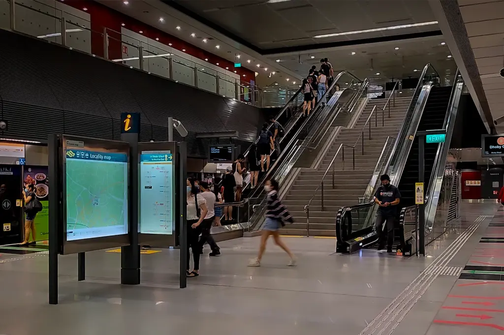 Interior view of Holland Village MRT Station in Singapore showing escalators, platform screen doors, station signage and commuters on the Circle Line platform area
