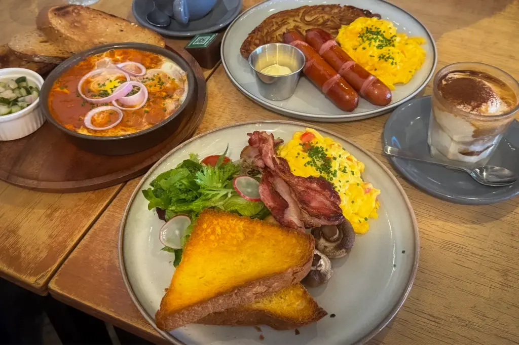 Brunch spread at Tolido’s Espresso Nook Singapore featuring shakshuka with toast, scrambled eggs with sausages and bacon, salad, mushrooms, cappuccino, and layered iced coffee on a wooden cafe table