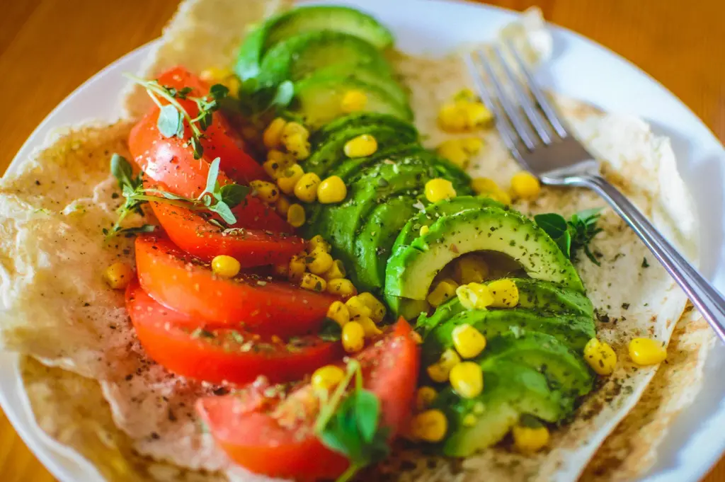 Fresh vegan wrap topped with sliced avocado, tomatoes, sweet corn, and herbs served on a white plate with a fork on a wooden table