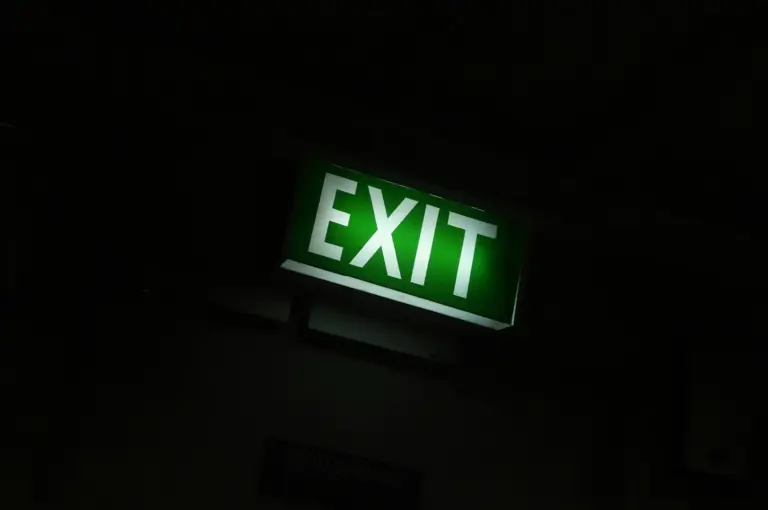 Illuminated green EXIT sign glowing in a dark indoor environment, mounted above an emergency exit with surrounding shadows creating a dramatic low-light scene.