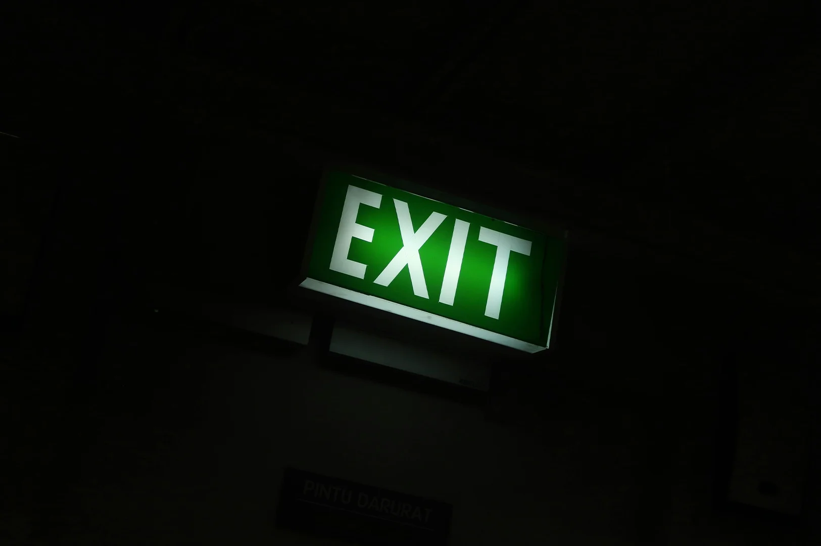 Illuminated green EXIT sign glowing in a dark indoor environment, mounted above an emergency exit with surrounding shadows creating a dramatic low-light scene.