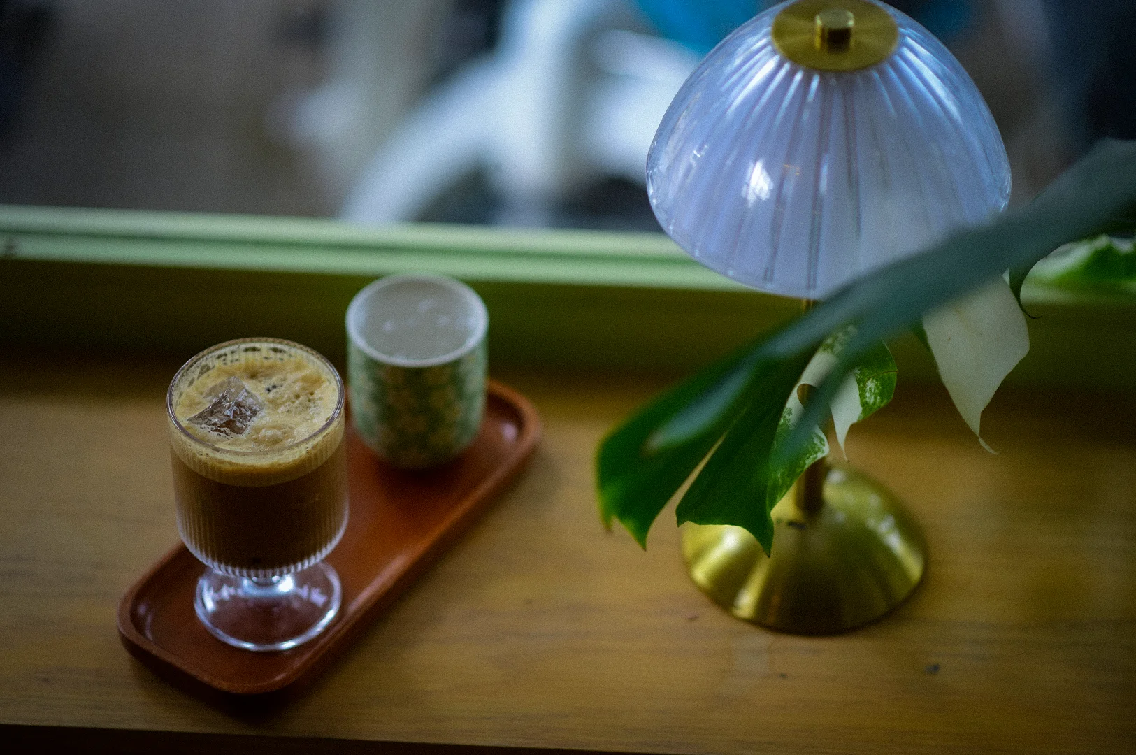 Iced coffee served on a wooden tray beside a decorative table lamp and leafy plant near a cafe window with soft natural lighting and cozy minimalist interior ambiance