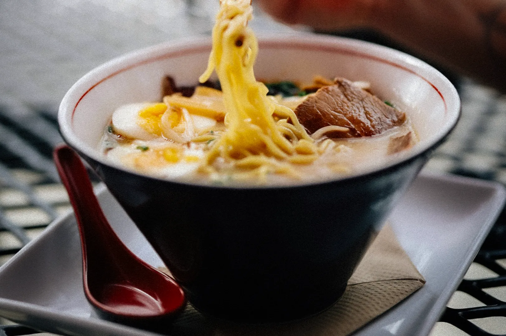 Close-up of a Japanese ramen bowl with noodles lifted by chopsticks, featuring sliced pork, soft-boiled egg, and rich broth. Warm food photography captures an authentic ramen dining experience at a restaurant table.