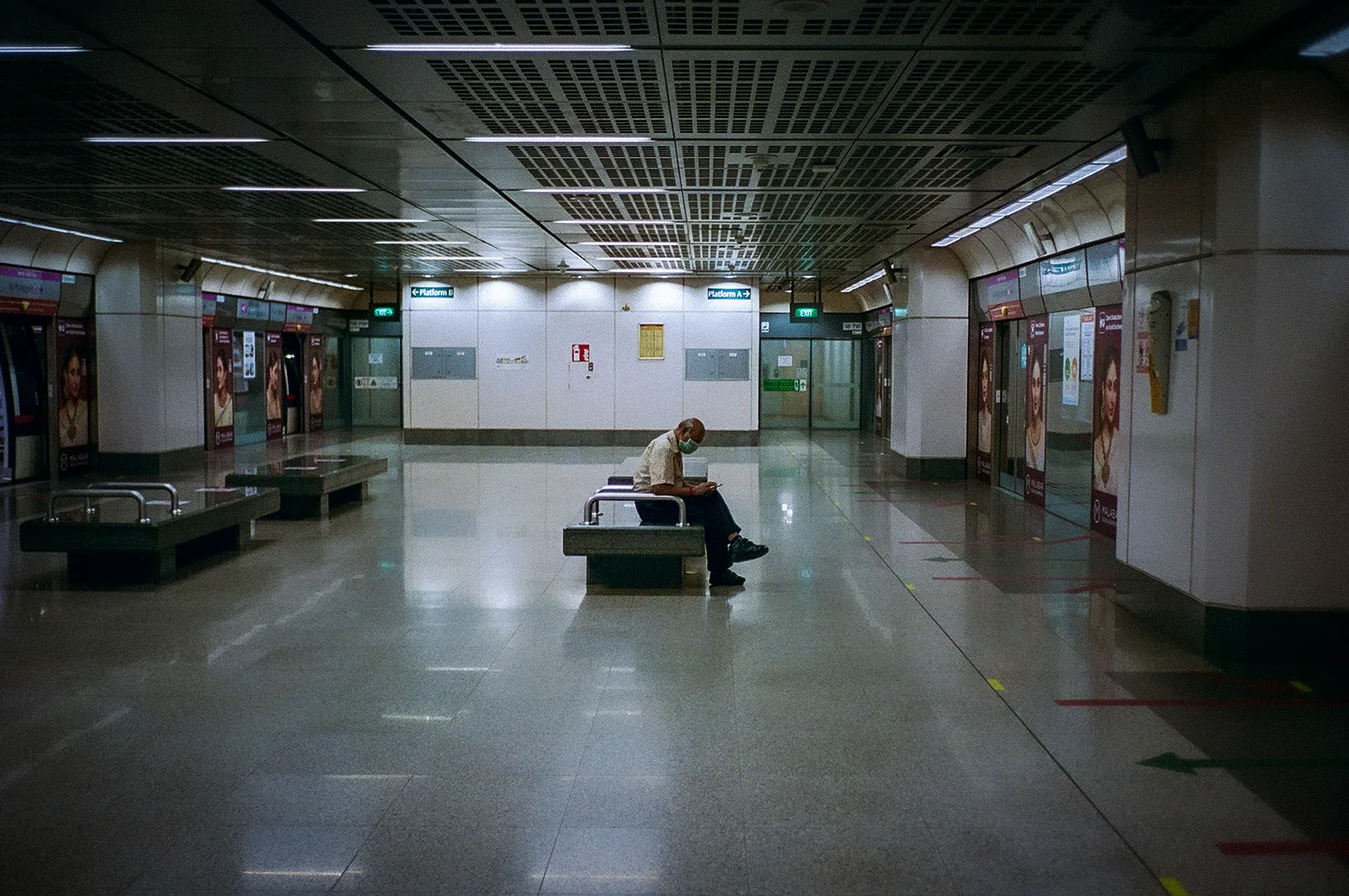Quiet Singapore MRT station platform with a lone commuter seated on a bench, surrounded by modern architecture, polished floors, platform screen doors, and overhead lighting.