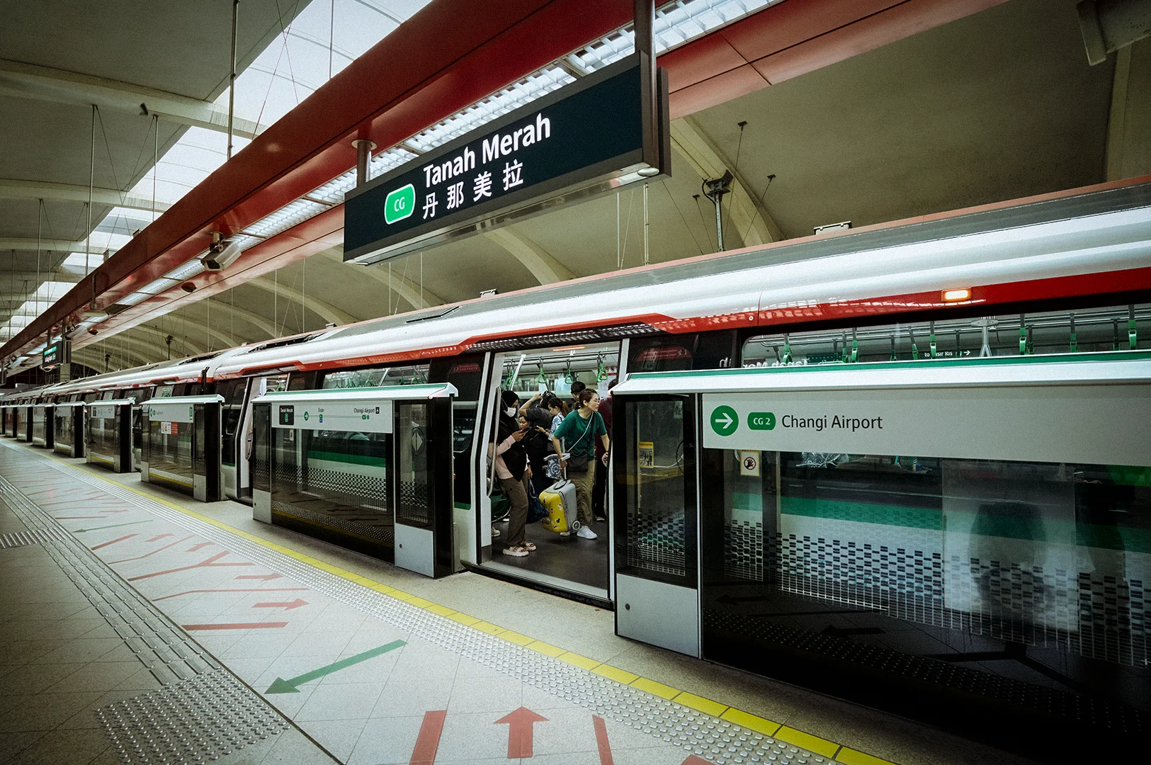 Passengers boarding an MRT train at Tanah Merah MRT Station in Singapore, with platform screen doors, station signage, and a train service heading toward Changi Airport on the East-West Line.