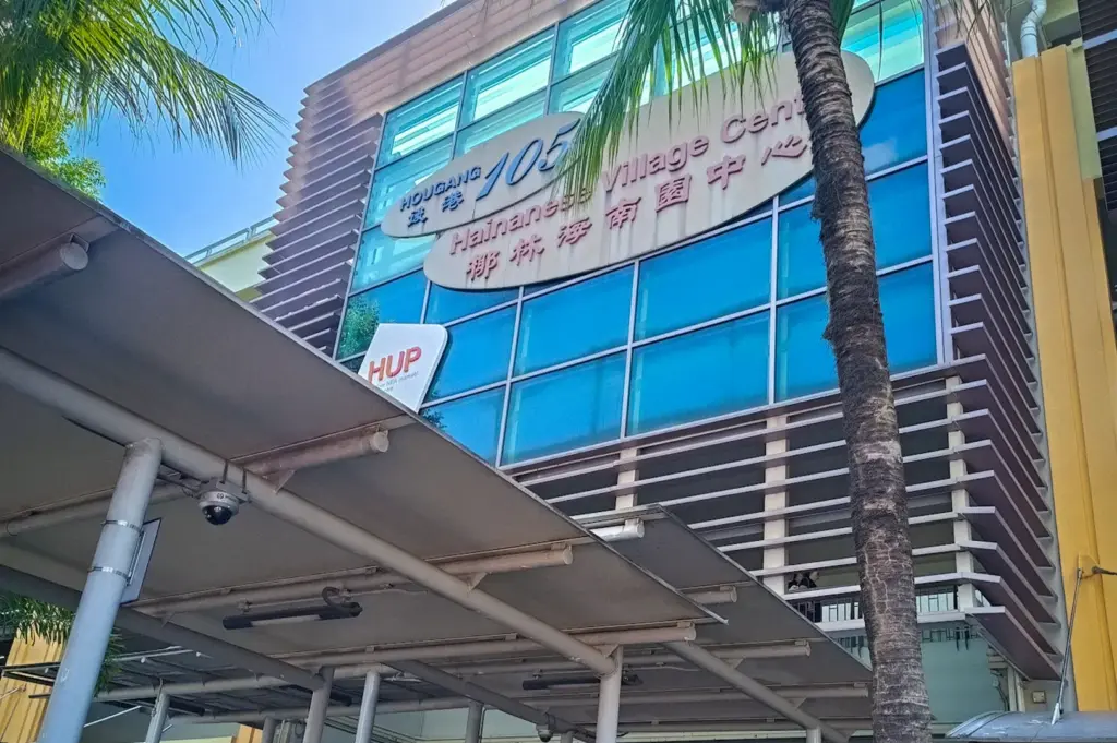 Exterior view of Hougang Village Centre in Singapore, featuring a multi-story commercial building with glass facade, covered pedestrian walkways, palm trees, escalators, and people entering and exiting the neighborhood shopping and food hub.