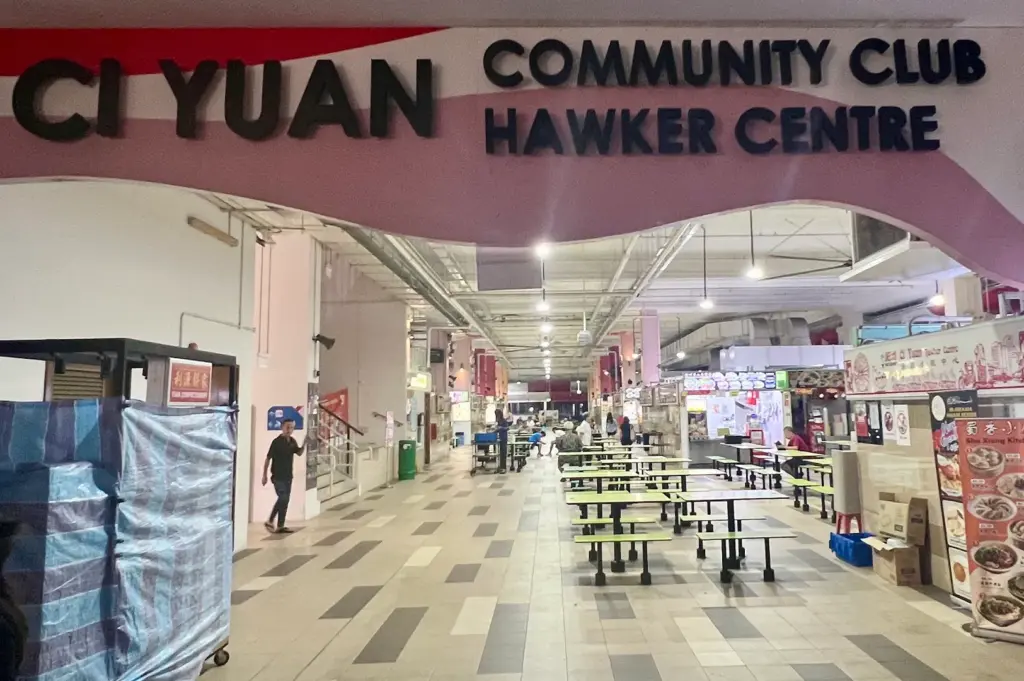 Entrance to Ci Yuan Community Club Hawker Centre in Singapore, featuring a covered food court with communal dining tables, local food stalls, illuminated signage, tiled flooring, and an open-air community dining environment.