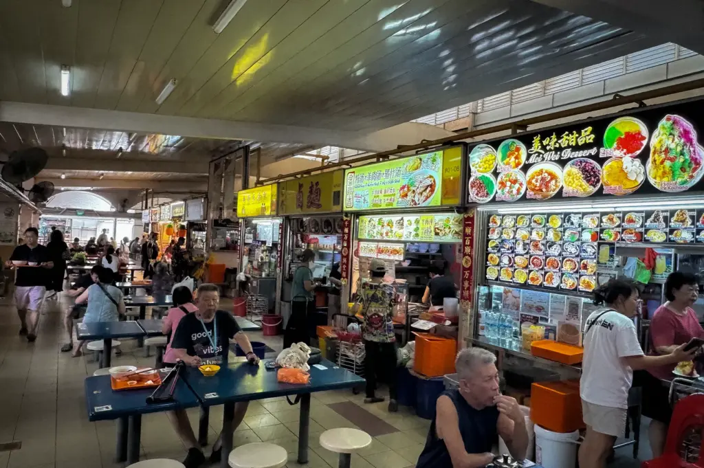 Interior of Kovan Market and Food Centre in Singapore, showing a bustling hawker centre with diners seated at communal tables, food stalls displaying colorful menu boards, and vendors serving a variety of local dishes in a traditional neighborhood food market setting.