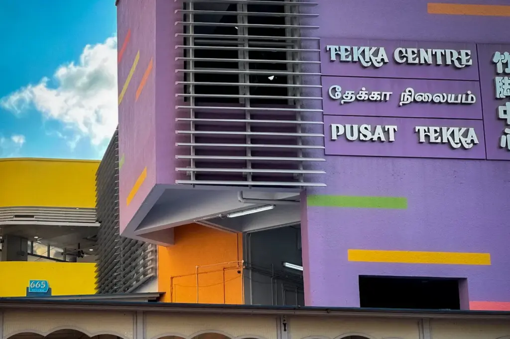 Exterior of Tekka Centre in Singapore's Little India district, featuring the building's distinctive purple and yellow facade with multilingual signage reading "Tekka Centre" and "Pusat Tekka." Pedestrians walk near the covered entrance to the popular hawker centre, wet market, and shopping complex.