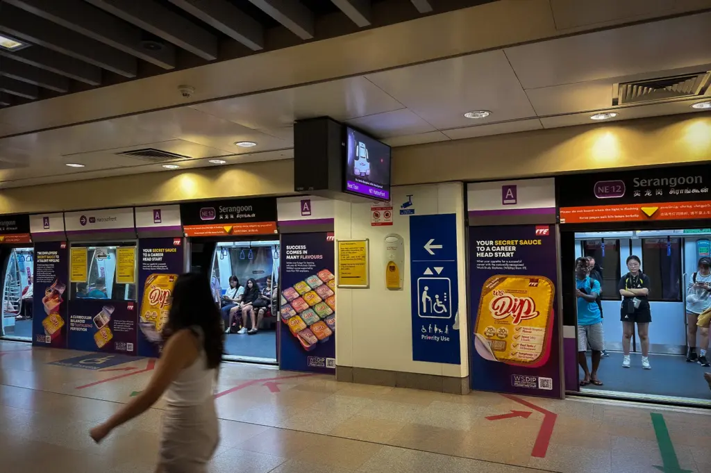 Platform of Singapore's North East Line MRT at Serangoon Station (NE12), showing a train stopped behind platform screen doors. Commuters wait to board and alight, while directional floor markings, priority-use signage, and station wayfinding signs are visible throughout the modern underground station.