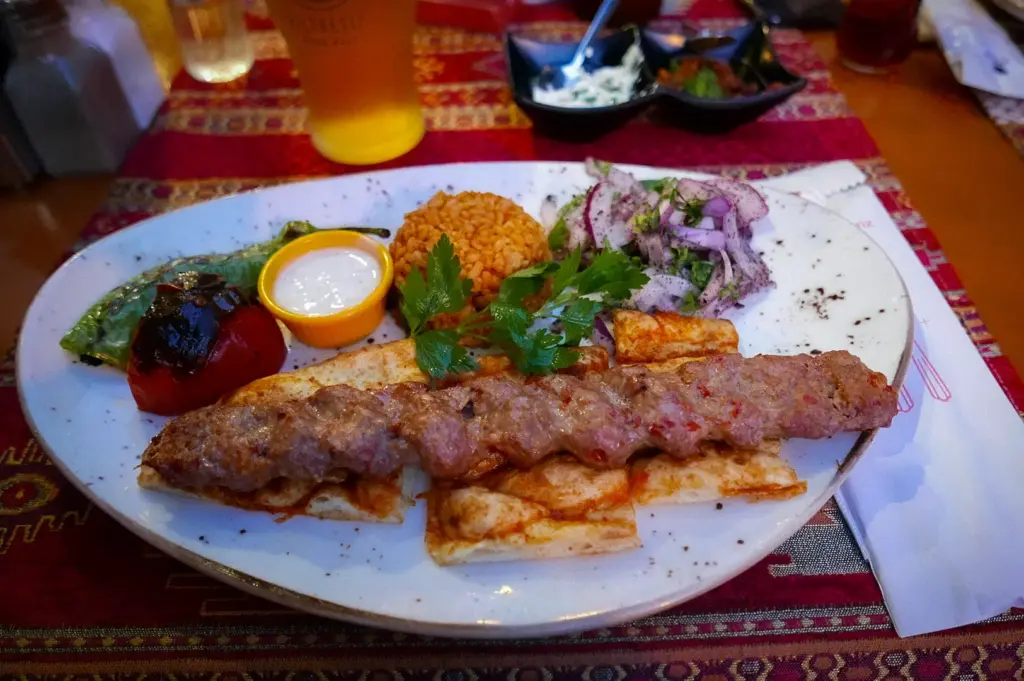 Halal Turkish kebab platter with grilled minced meat skewer, rice, pita bread, yogurt sauce, roasted vegetables, and fresh onion salad served at a restaurant.