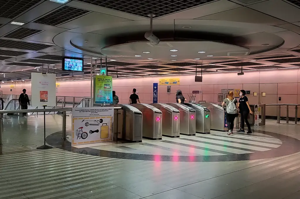 Kovan MRT Station concourse in Singapore featuring fare gates, commuters entering and exiting, overhead signage, and a spacious underground transit hall.