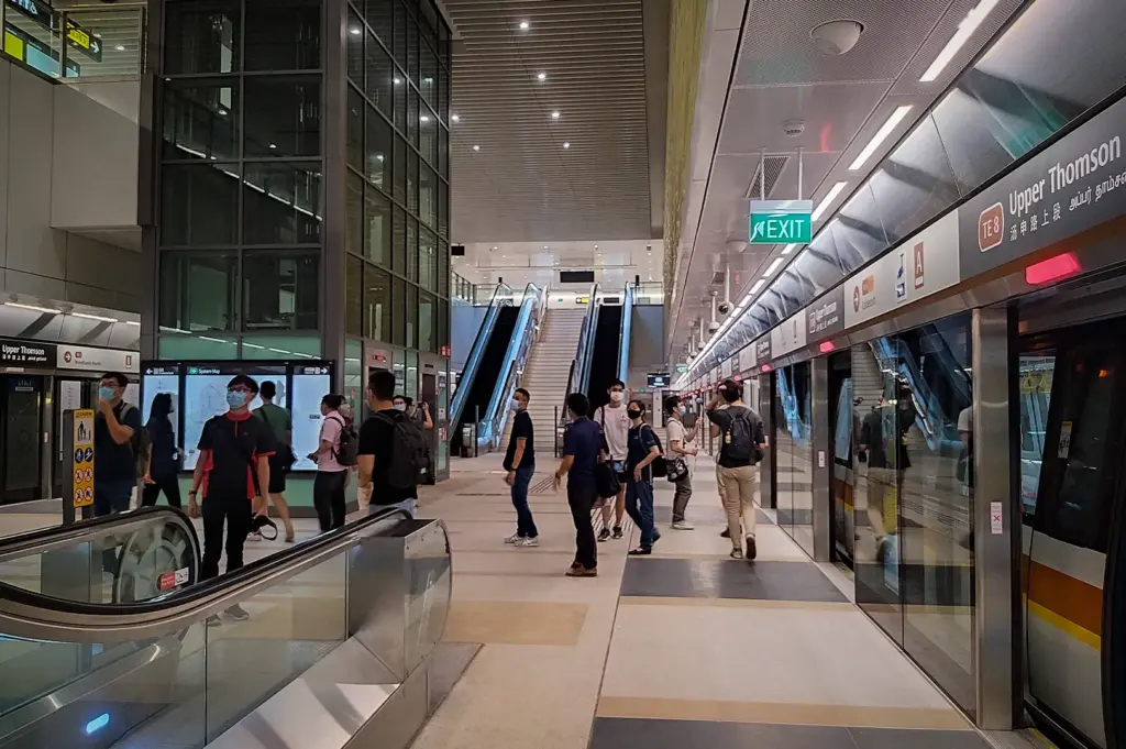 Interior view of Upper Thomson MRT Station in Singapore, showing the platform level with passengers waiting near train doors, escalators, glass elevators, platform screen doors, and modern station architecture on the Thomson-East Coast Line.