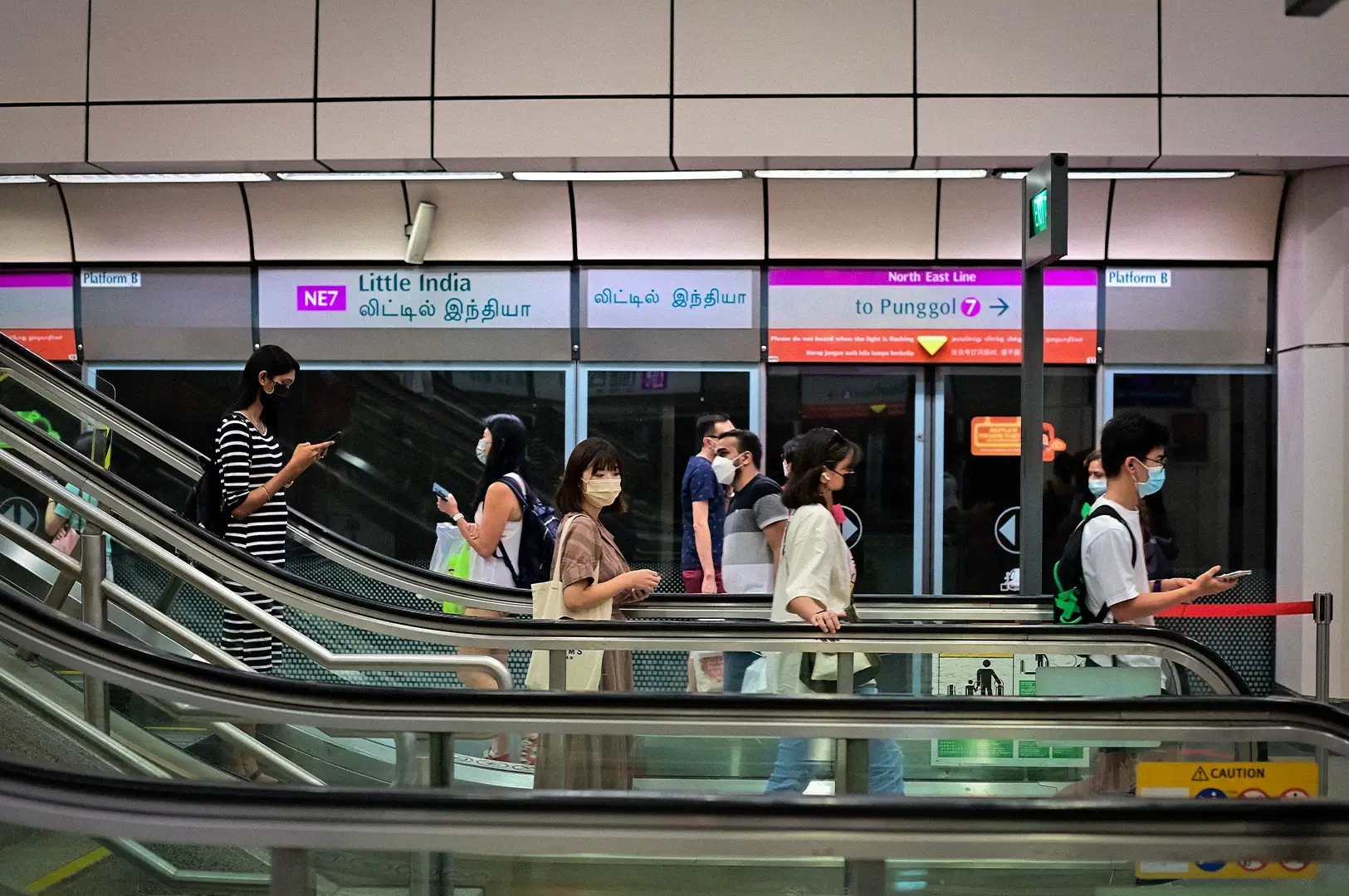 Commuters riding escalators inside Singapore MRT Little India Station with platform signage, train doors, and a busy public transport concourse.