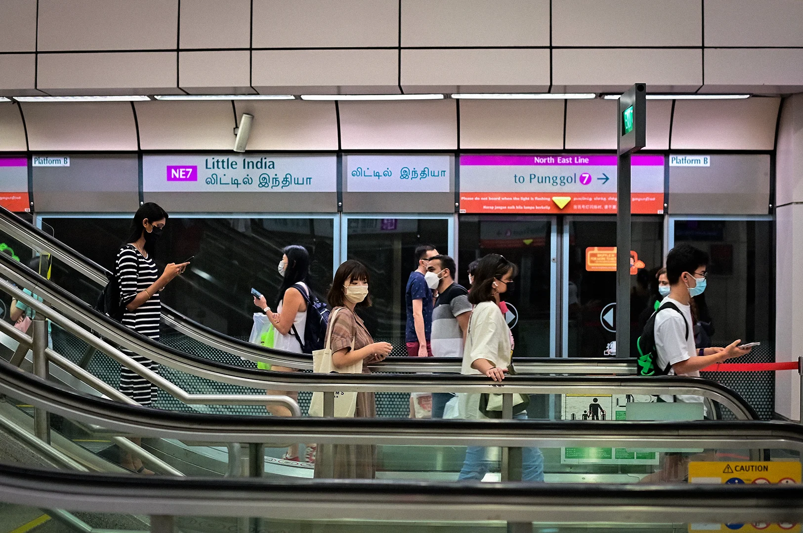 Commuters riding escalators inside Little India MRT Station in Singapore with platform screen doors, station signage, and modern public transport infrastructure.